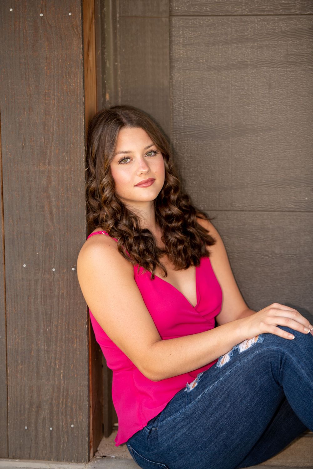 Portrait of someone in a pink top and jeans sitting casually against a wooden wall.
