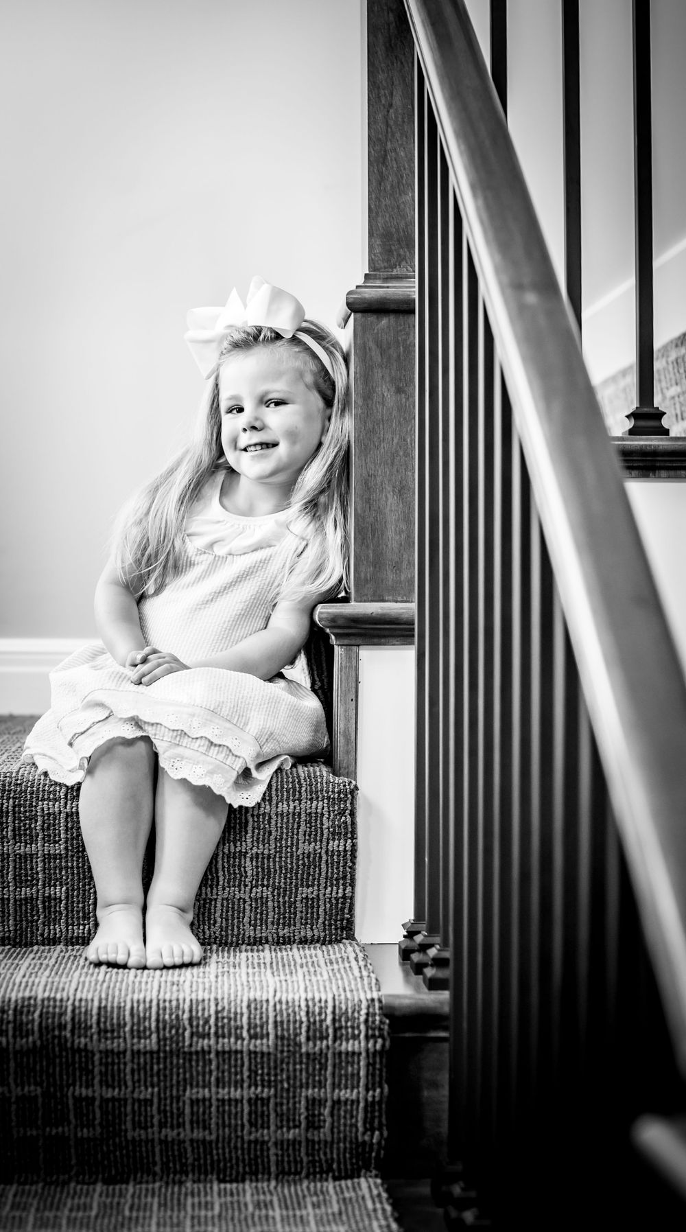 Black and white portrait of a child sitting on carpeted stairs with wooden banister.