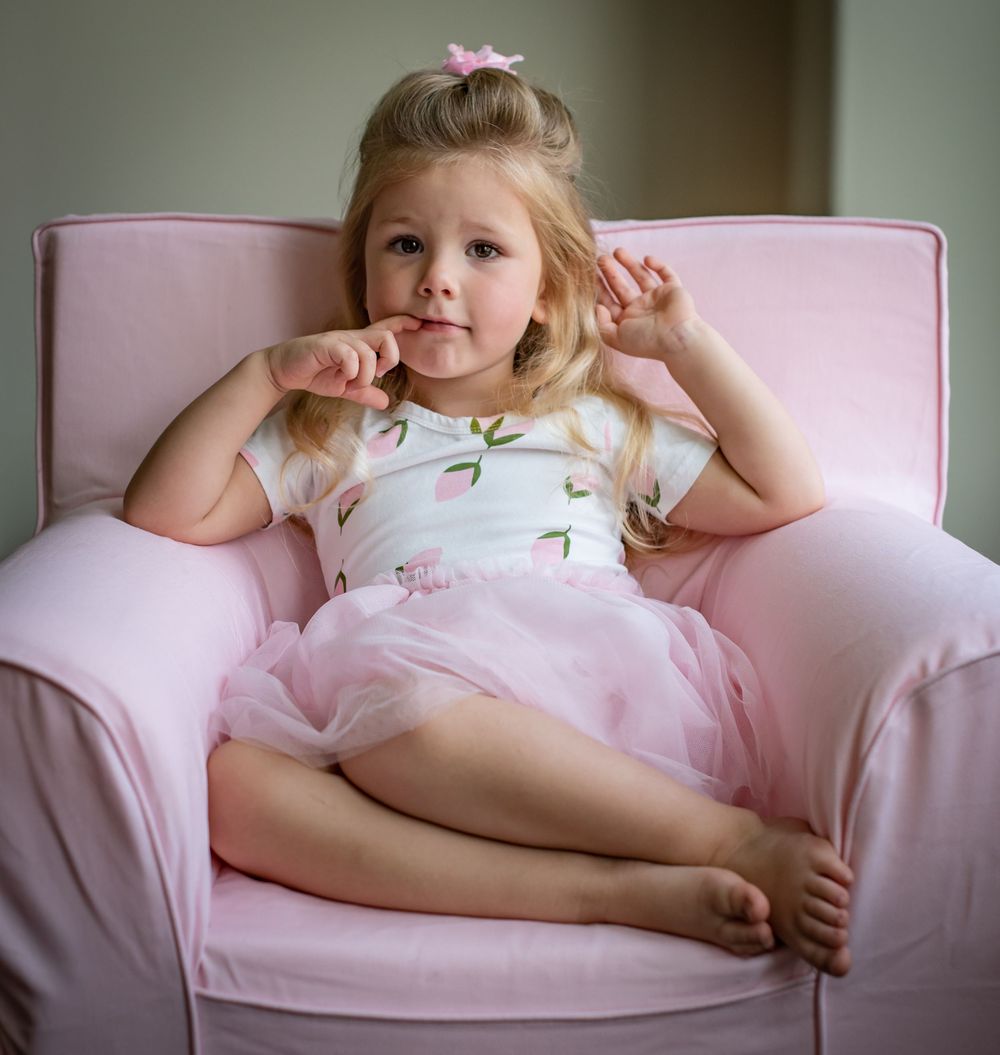 Child relaxing in pink armchair wearing tulle dress and making playful expression.