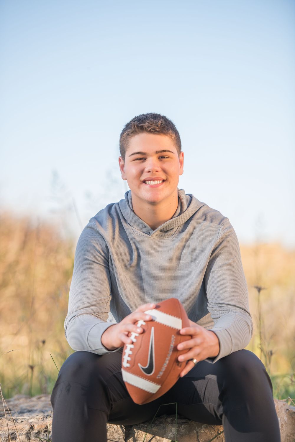 Athletic portrait of someone holding football while sitting outdoors in grey hoodie.