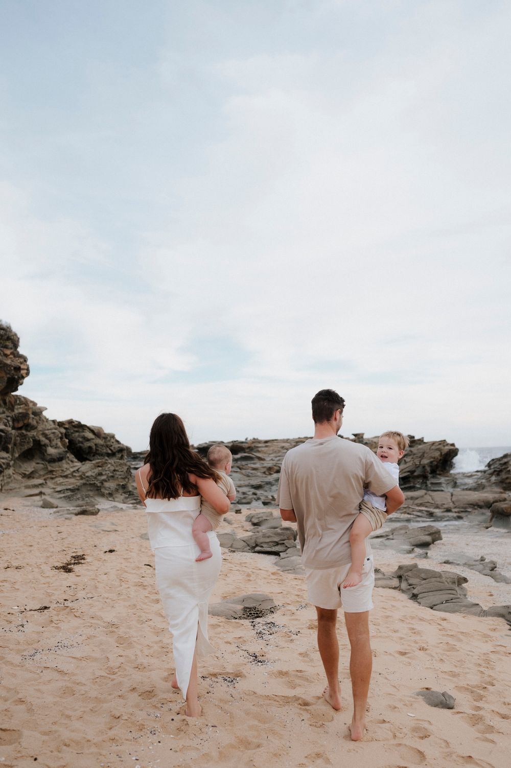 Family walking barefoot on rocky beach shore at sunset.