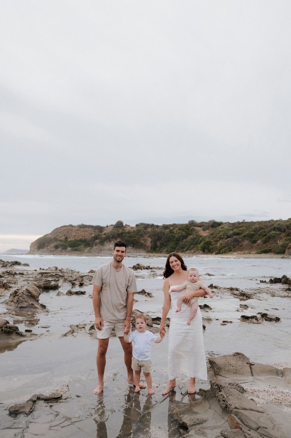 Family of four standing barefoot on rocky beach during cloudy day.
