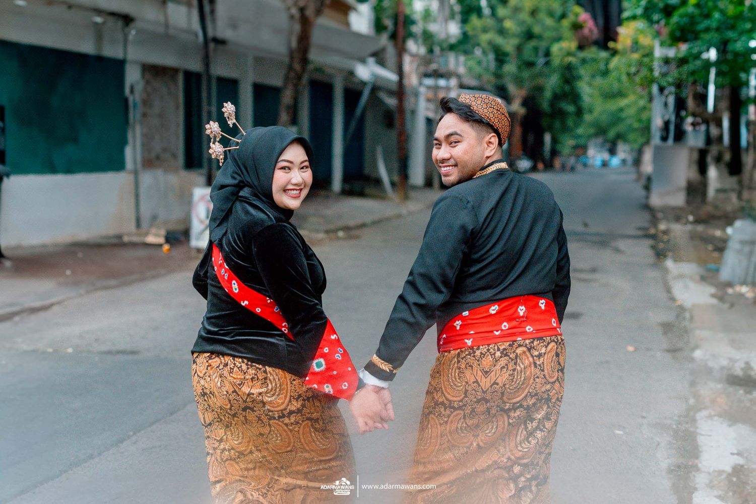 Couple wearing traditional Indonesian batik and dark tops walk hand in hand on a quiet street.