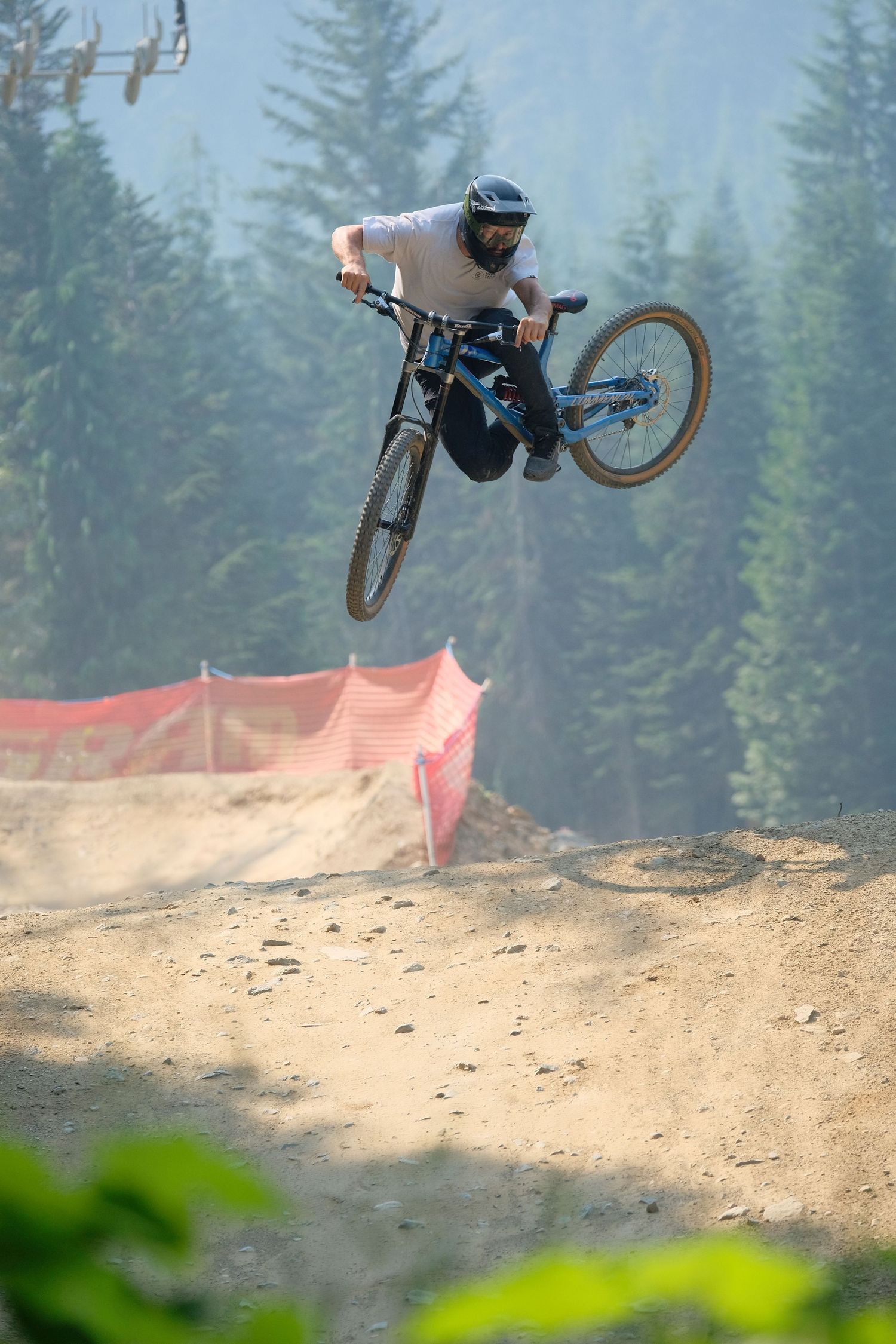A mountain biker performs a jump on a dirt track with evergreen trees and orange netting visible in the background.