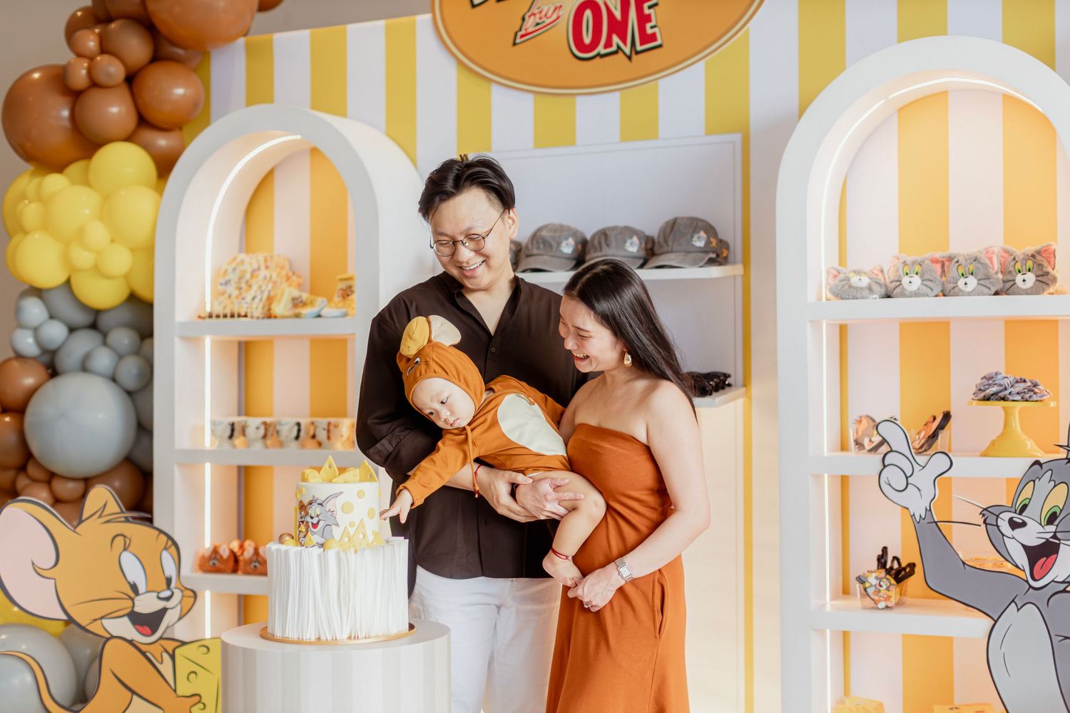 Couple admiring a Tom and Jerry themed first birthday cake at a yellow and orange decorated party.