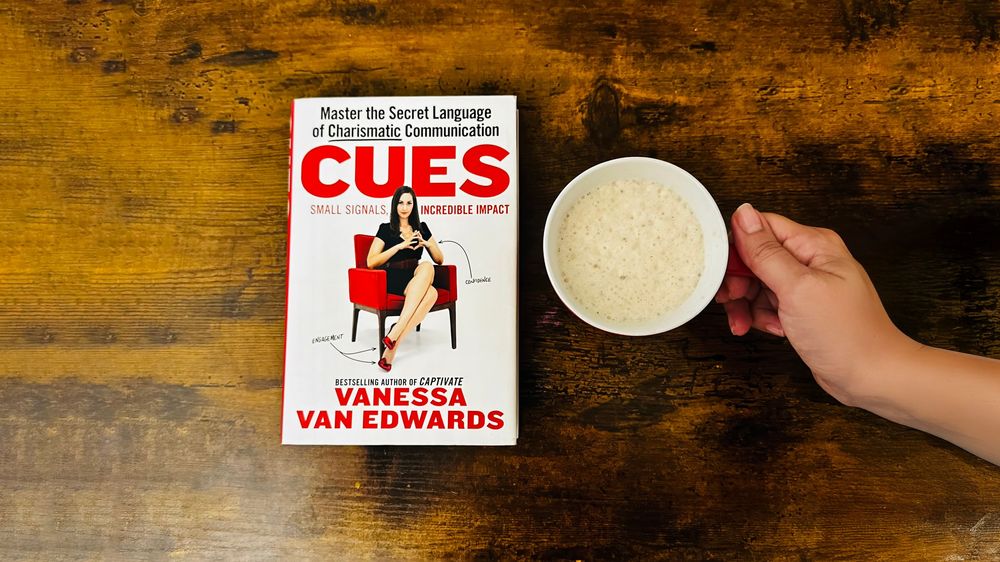 The book Cues, by Vanessa Van Edwards is on a table next to a cup of coffee.
