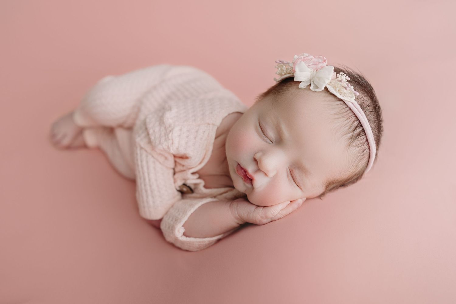 Sleeping newborn in pink knit outfit and floral headband rests peacefully on rosy backdrop during professional photoshoot.