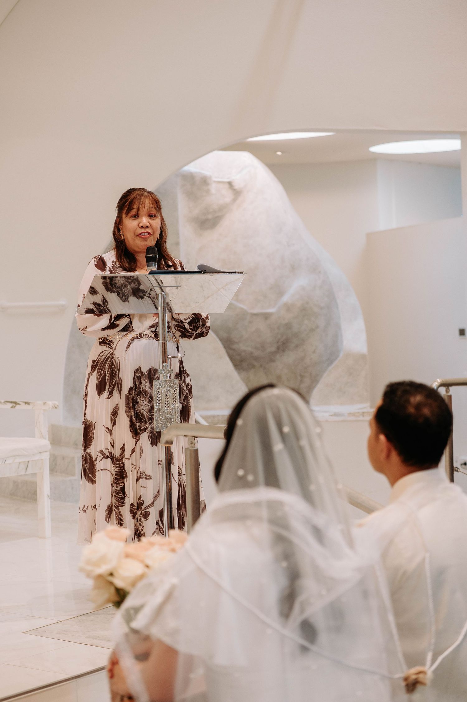 A person at a podium speaking during a wedding ceremony in an all-white modern space with a bride and groom seated.