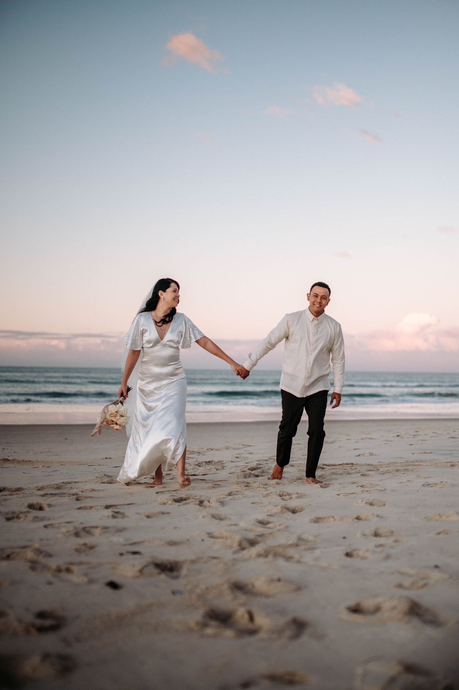 A couple in white clothing walk hand in hand on a sandy beach at sunset with gentle ocean waves in the background.