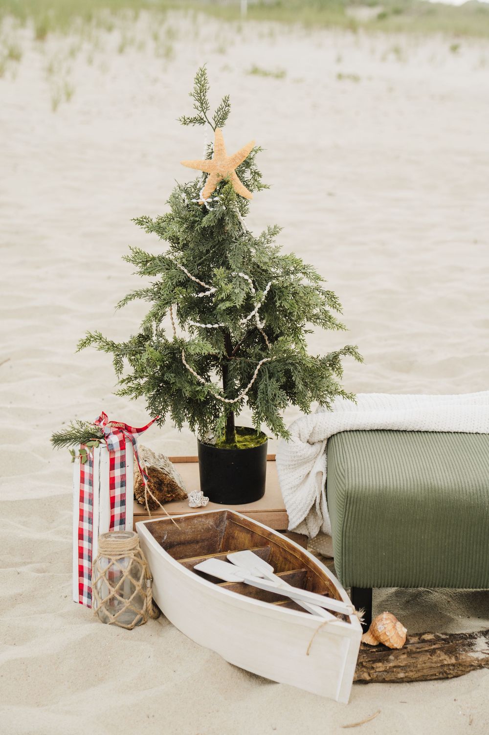 Beach Christmas scene with small decorated tree topped with starfish, white rowboat, lantern, and green ottoman on sand.