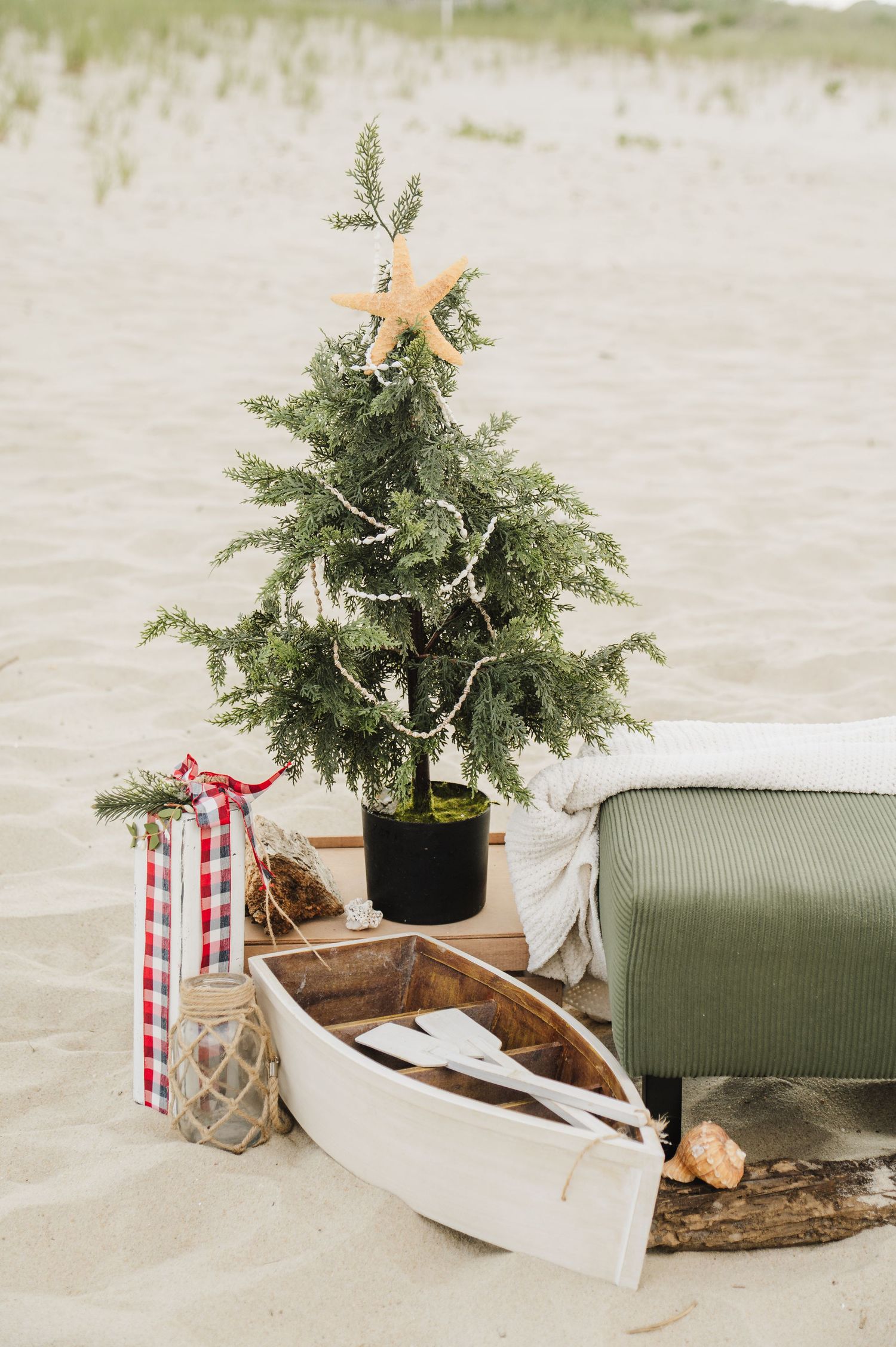 Beach Christmas scene with small decorated tree topped with starfish, white rowboat, lantern, and green ottoman on sand.