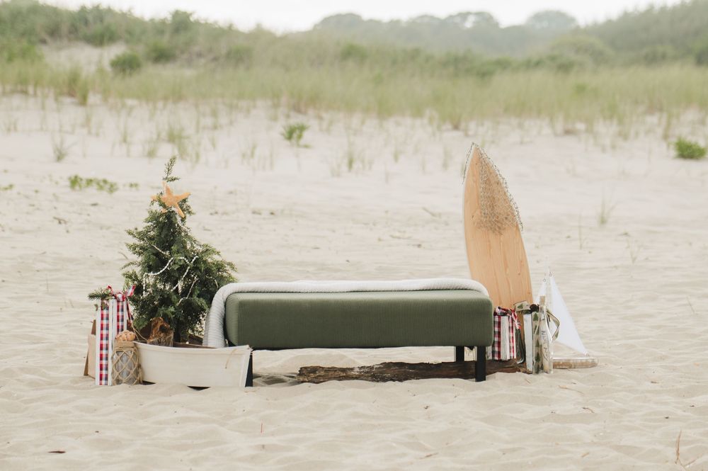 Beach Christmas scene with green bench, surfboard, decorated tree, and wrapped gifts on sandy shore.