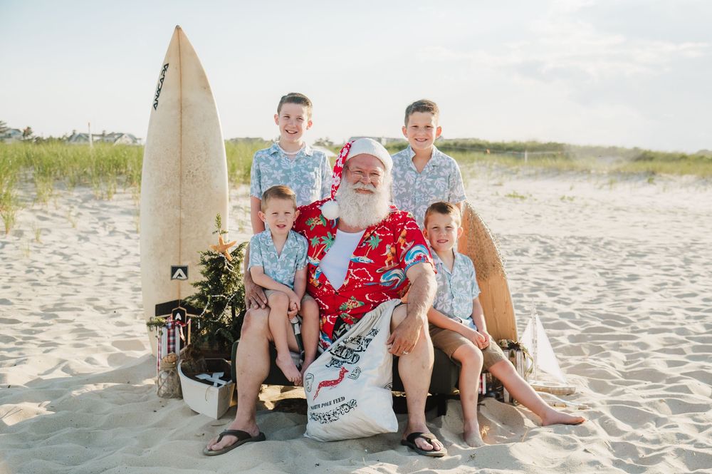 Four boys pose with Santa in a red Hawaiian shirt on a sunny beach with a surfboard.