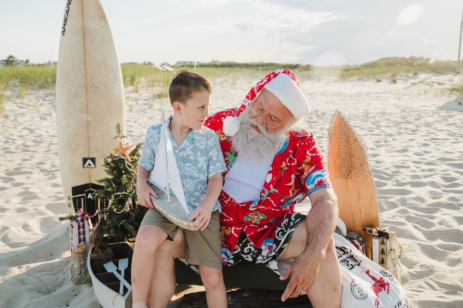A young boy sits with Santa Claus in a red Hawaiian shirt on a beach, sharing a warm moment.