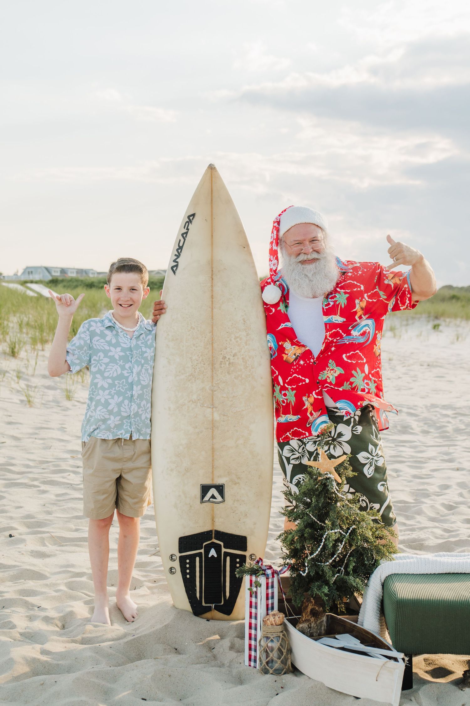 A boy and beach Santa give shaka signs beside a surfboard and small Christmas tree on sand.