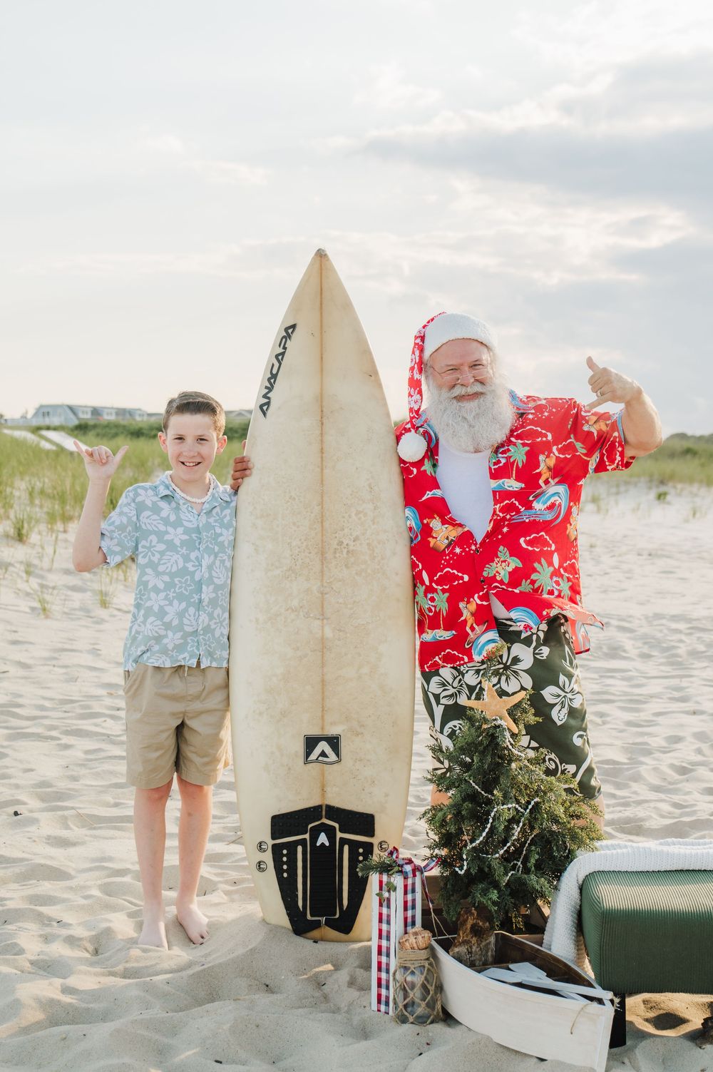 A boy and beach Santa give shaka signs beside a surfboard and small Christmas tree on sand.