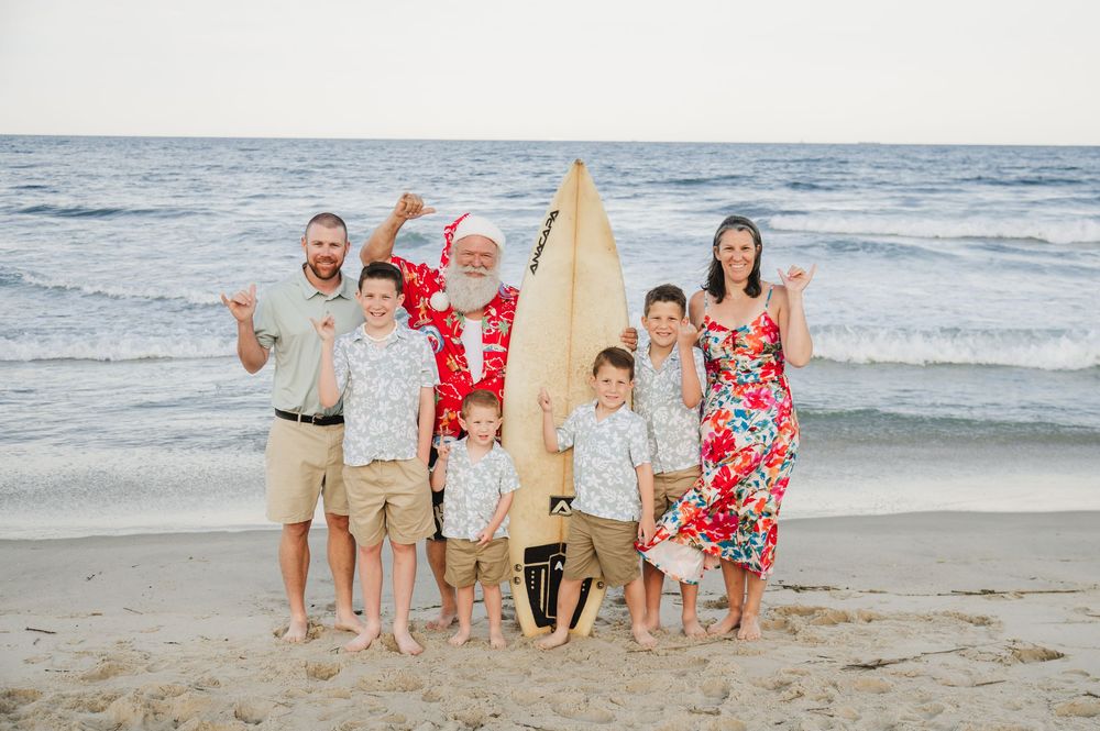 Family posing with Santa in Hawaiian shirt holding surfboard on sunny beach holiday photo.