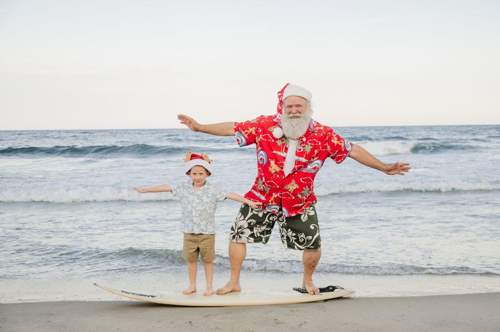 Santa Claus and child in Christmas hats surf together on a sunny beach wearing festive Hawaiian outfits.