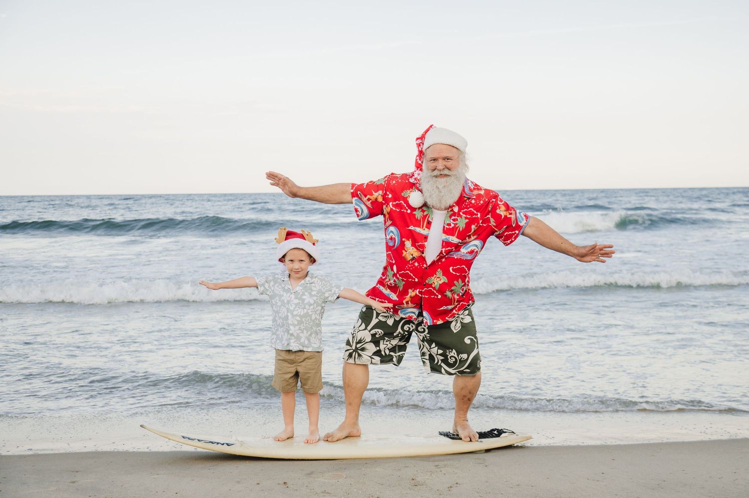 Santa Claus and child in Christmas hats surf together on a sunny beach wearing festive Hawaiian outfits.