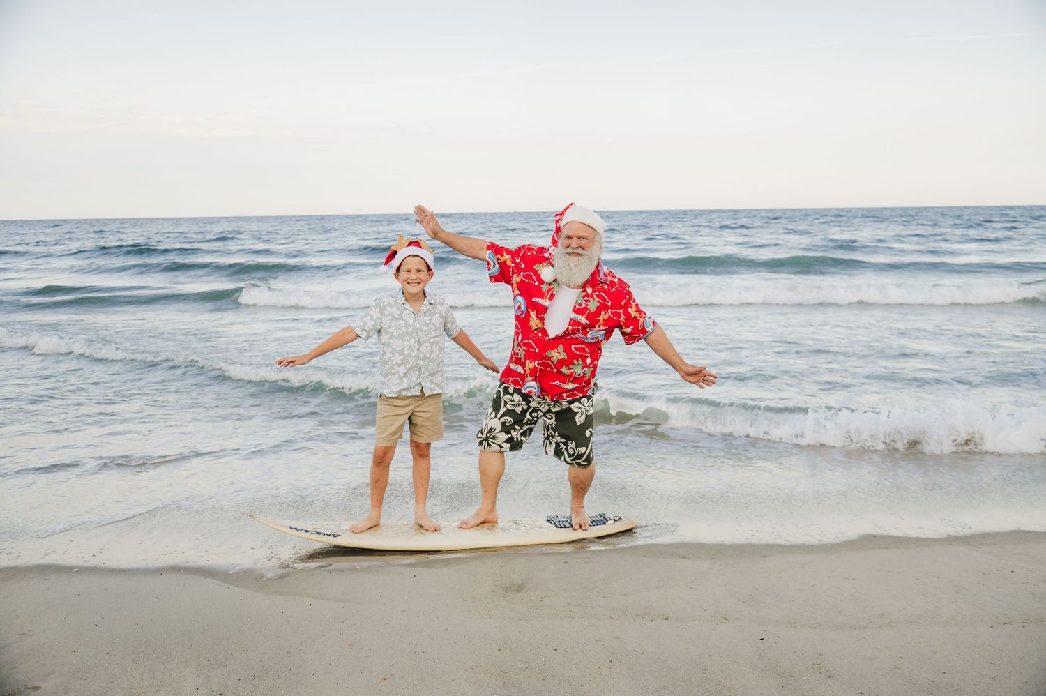Two joyful people in Santa hats celebrate Christmas on a sunny beach, balancing on a surfboard near ocean waves.