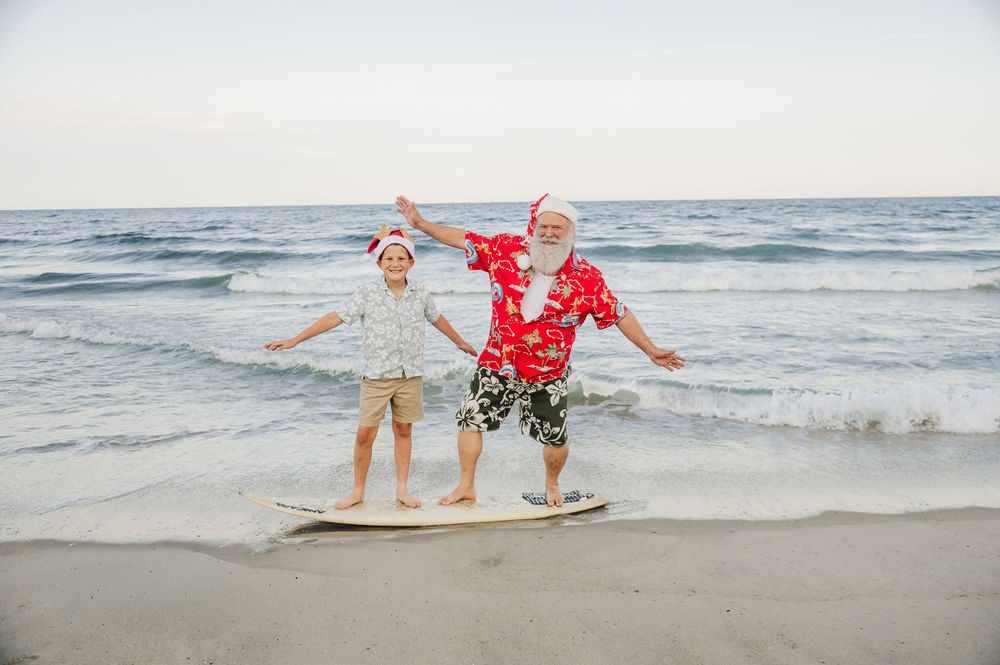 Two joyful people in Santa hats celebrate Christmas on a sunny beach, balancing on a surfboard near ocean waves.