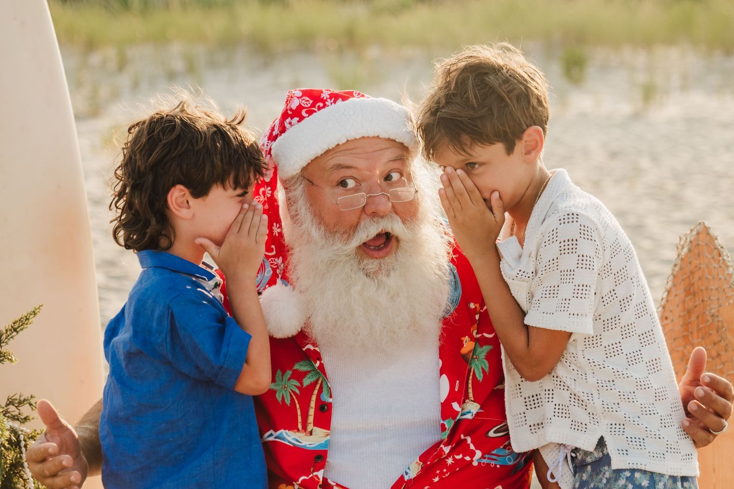 Two children whisper secrets to Santa Claus in a Hawaiian shirt on a sunny beach holiday.