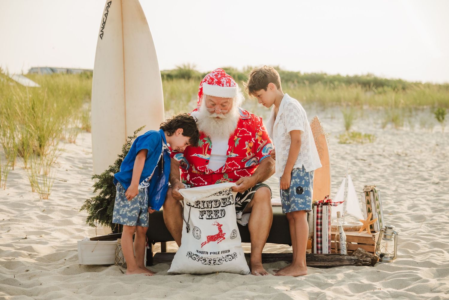 Santa Claus in Hawaiian shirt with two boys opening gift sack on sandy beach with surfboard at Christmas.