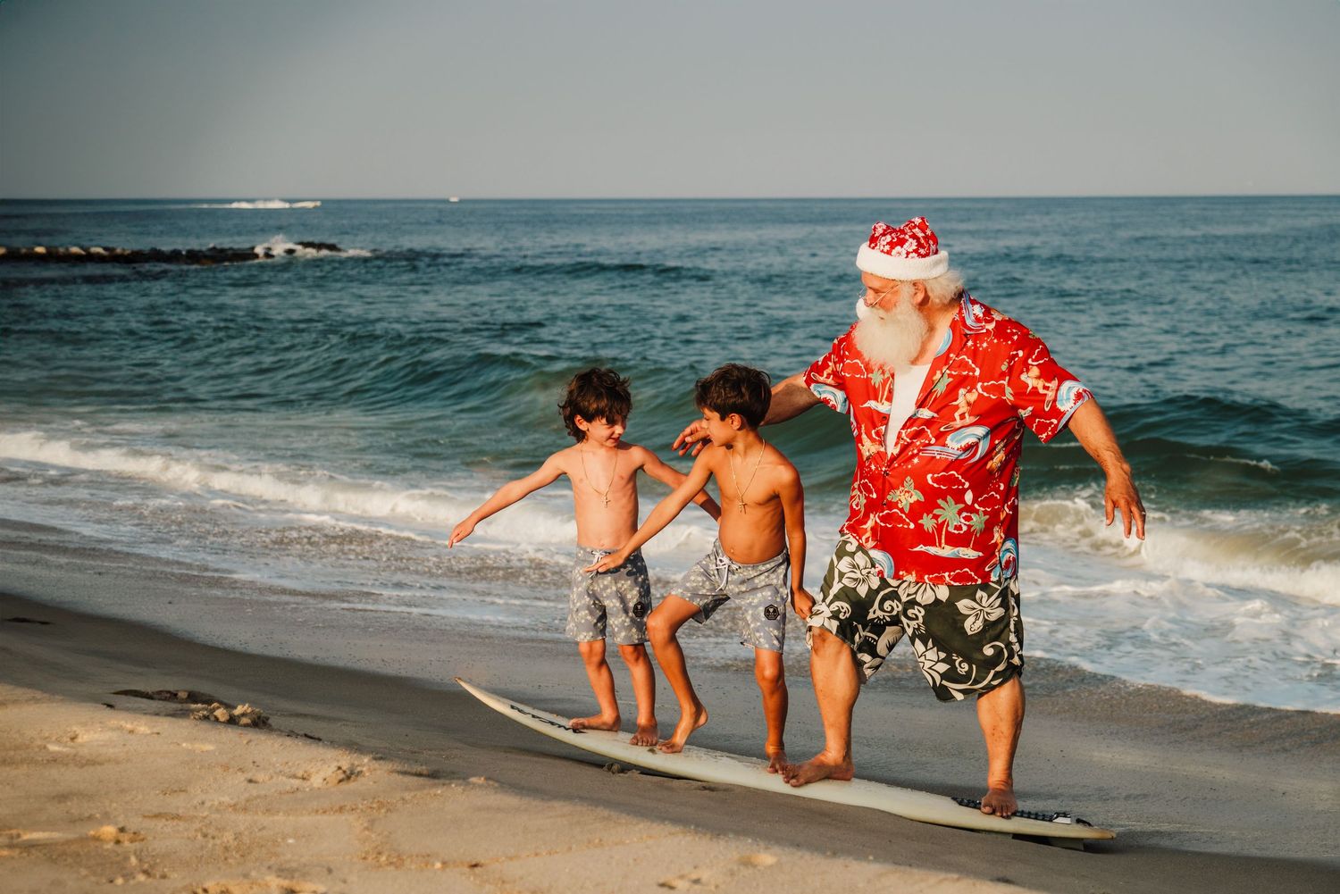 Santa Claus in Hawaiian shirt teaches two children to surf on a sunny beach holiday.