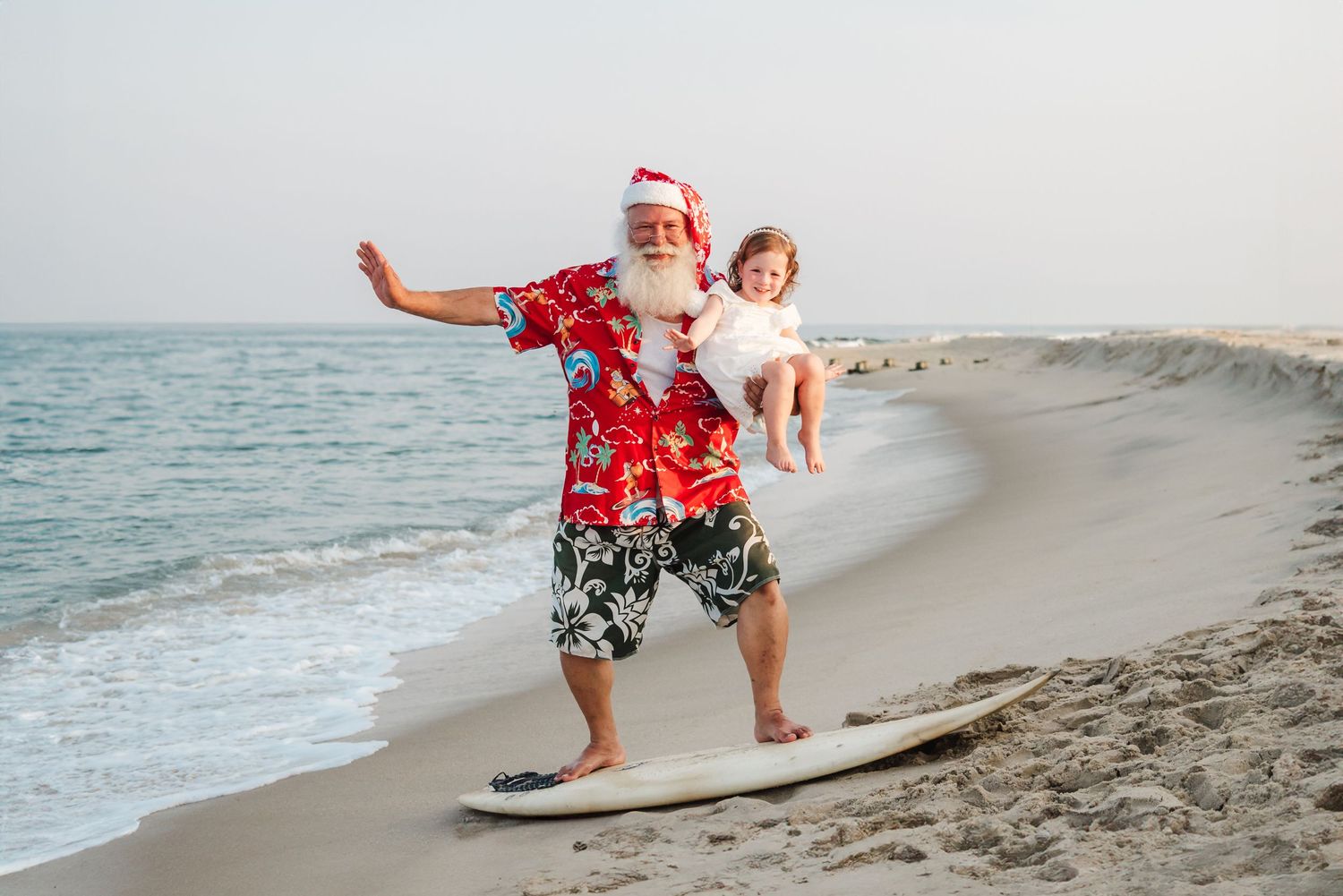 Santa Claus in Hawaiian shirt holds baby on surfboard at sunny beach, blending Christmas and tropical vacation themes.