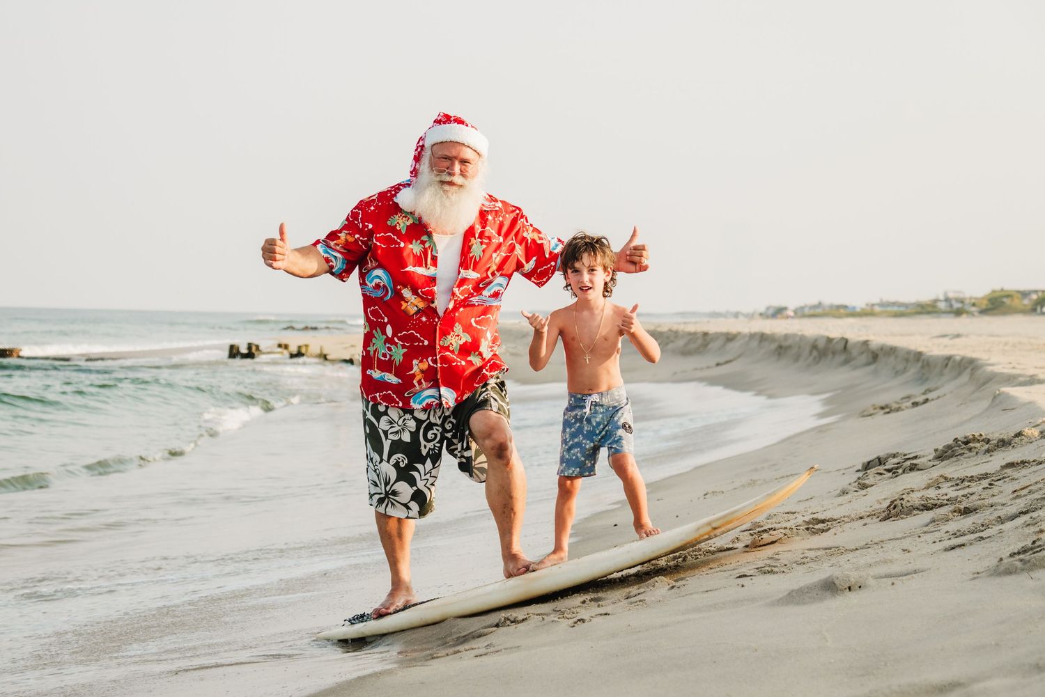 Santa Claus in Hawaiian shirt surfs with a young boy on a sunny beach, both smiling and giving thumbs up.