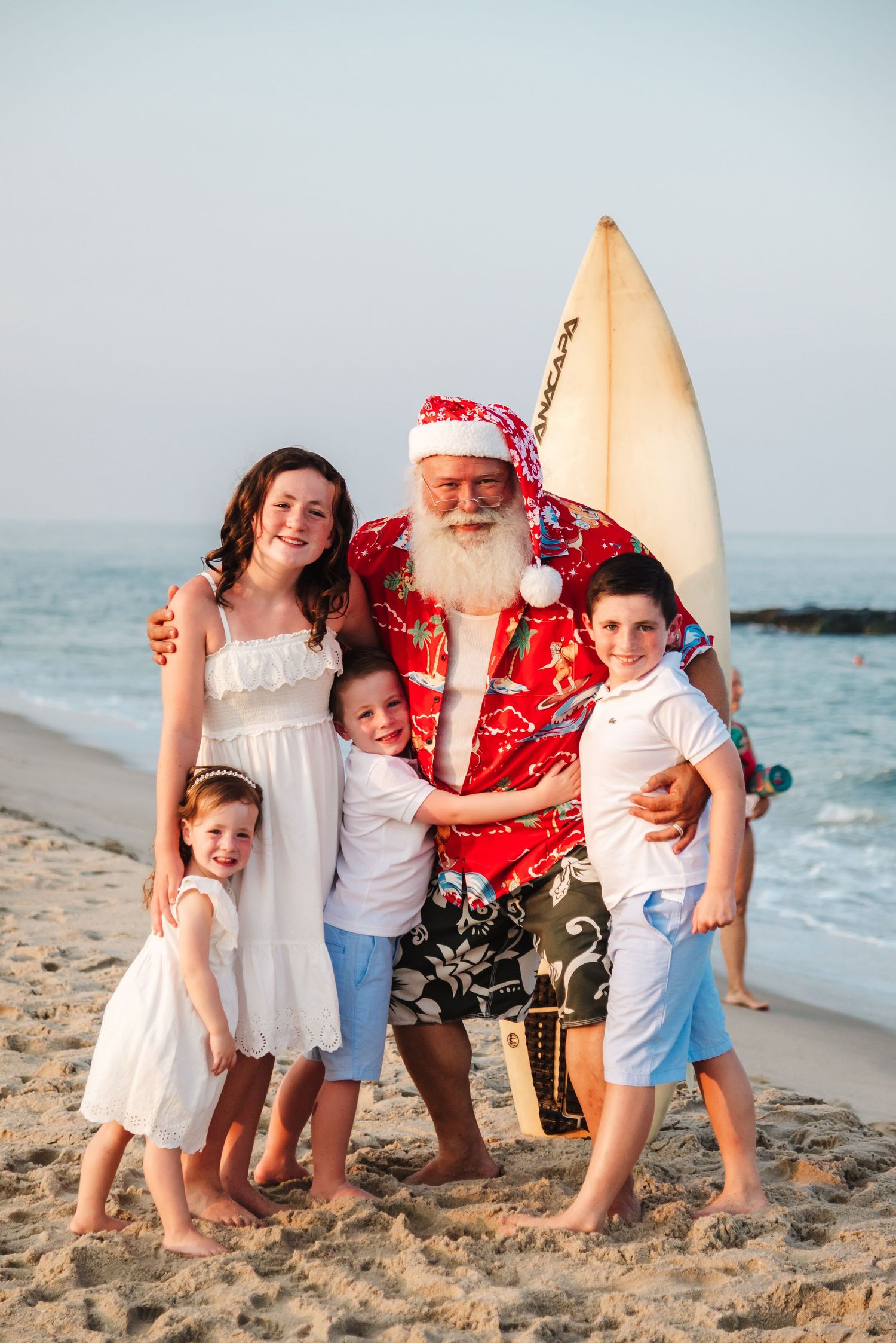 Santa Claus in Hawaiian shirt holds surfboard while posing with three children on a sunny beach.