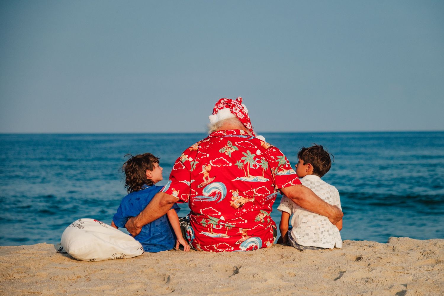 Santa Claus in a red Hawaiian shirt sits on a beach with two children, ocean in the background.