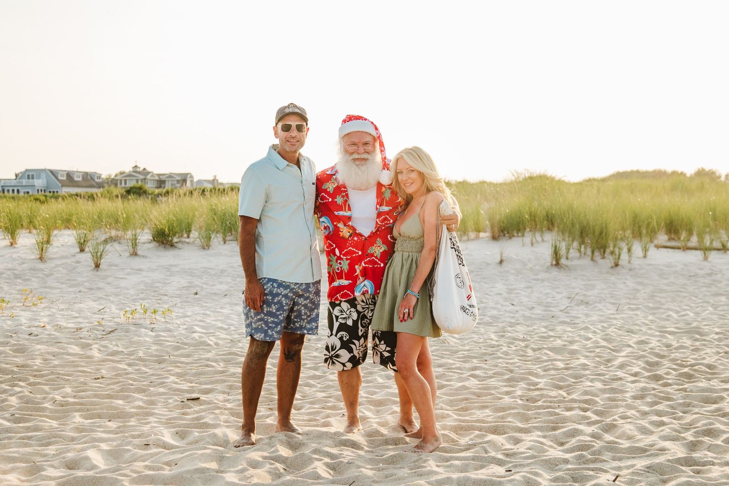 Couple posing with Santa Claus in festive beach attire on sandy shore with dunes in background.