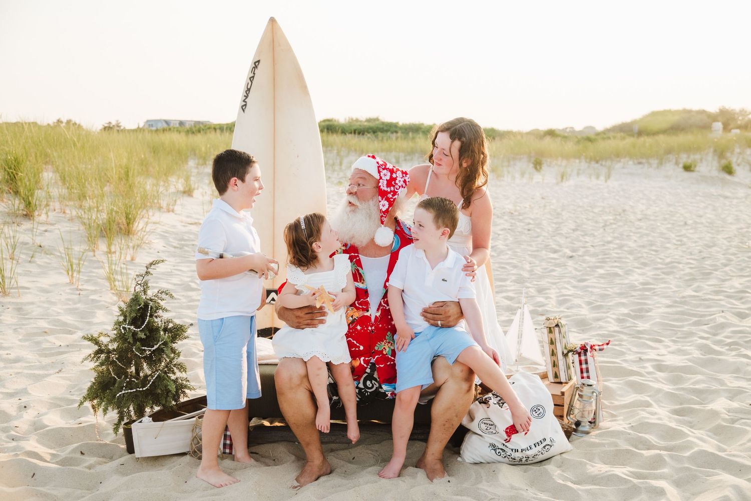 Family poses with Santa in red swim trunks on sandy beach with surfboard and small Christmas tree.