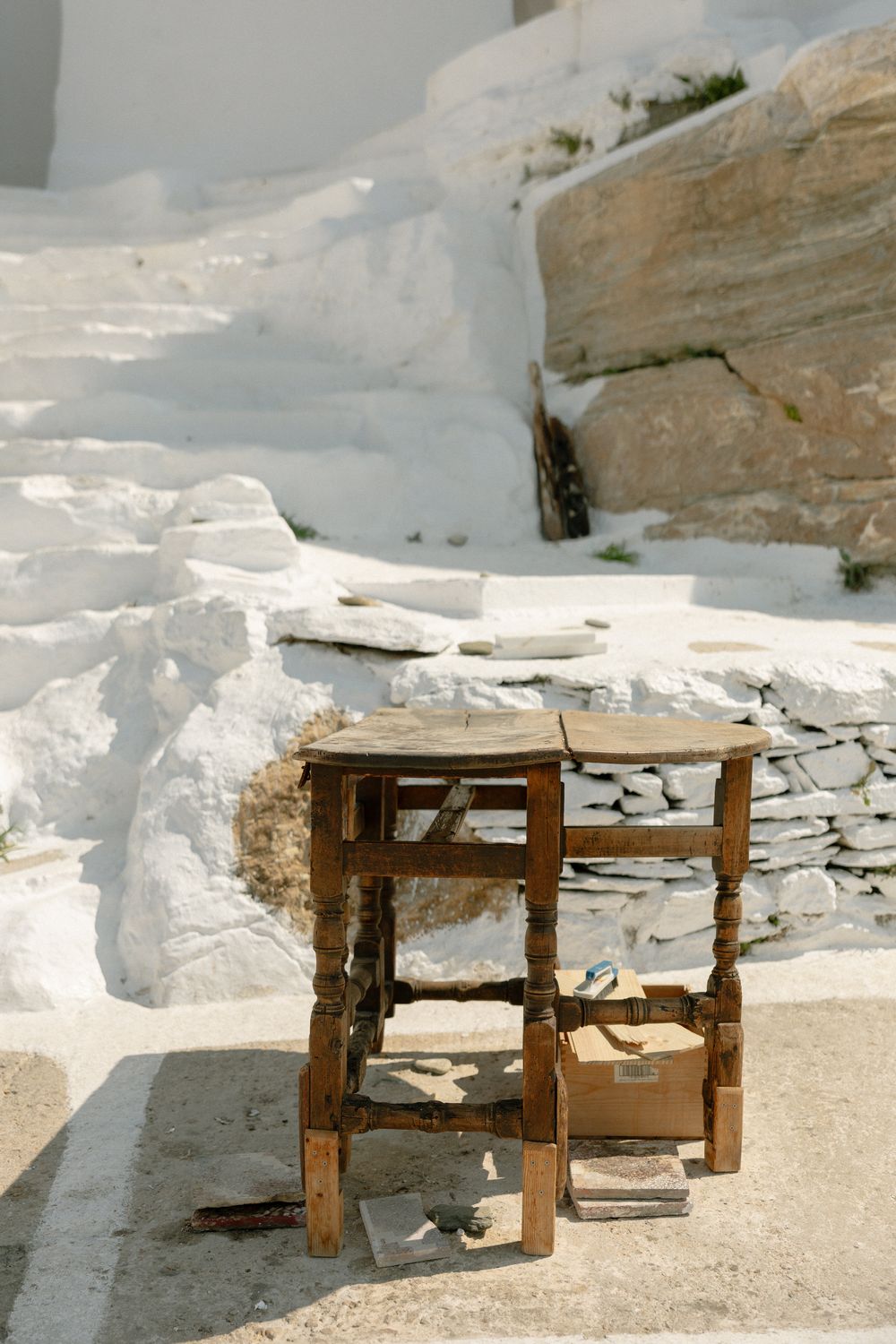 Rustic wooden stool sits against whitewashed stone wall with snow-covered steps in Mediterranean setting.