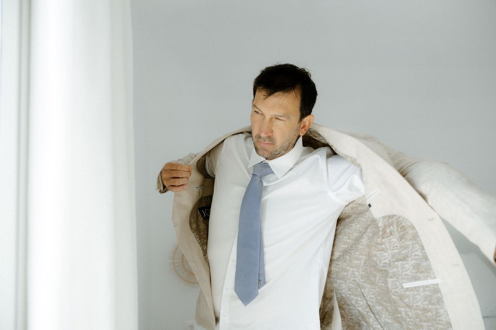 Person in light suit and blue tie adjusting clothing in minimalist white interior.