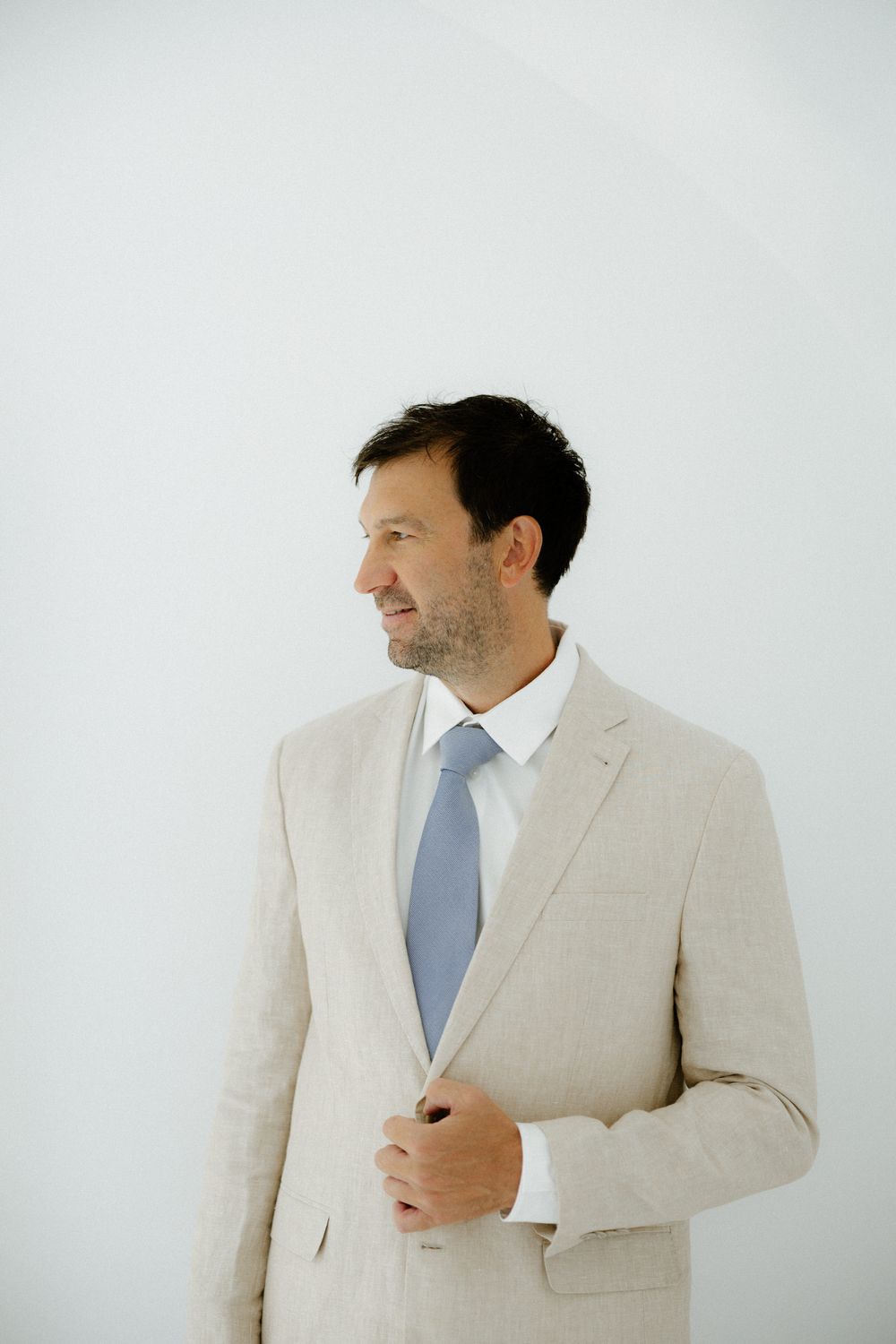 Person in beige suit and blue tie poses against white wall in natural light.