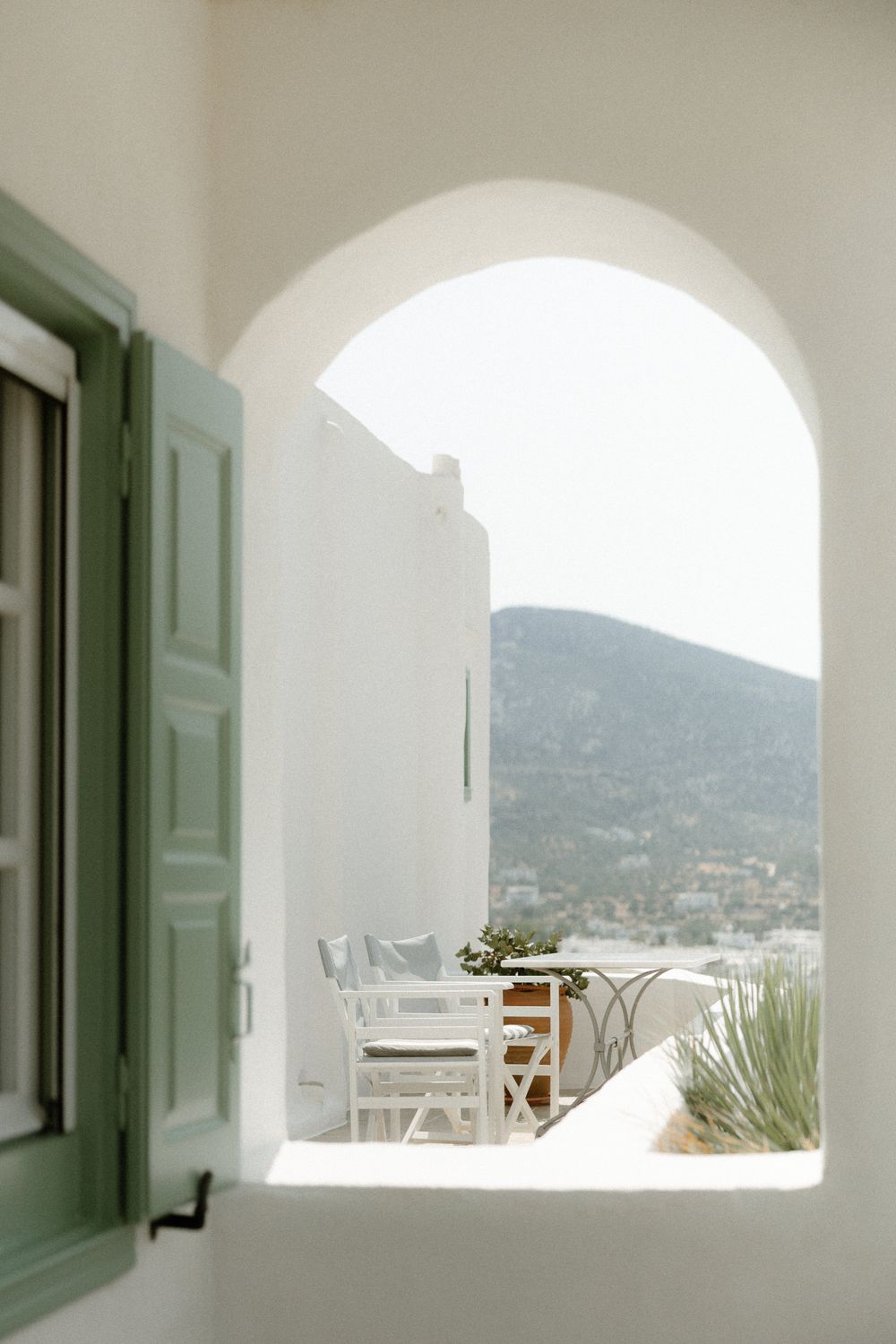 Arched window frames mountain view with white patio furniture and green shutters. Wedding photographer Sifnos