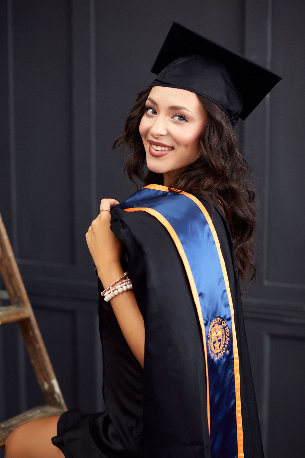 Graduate in black cap and gown with blue sash poses for celebratory portraits.