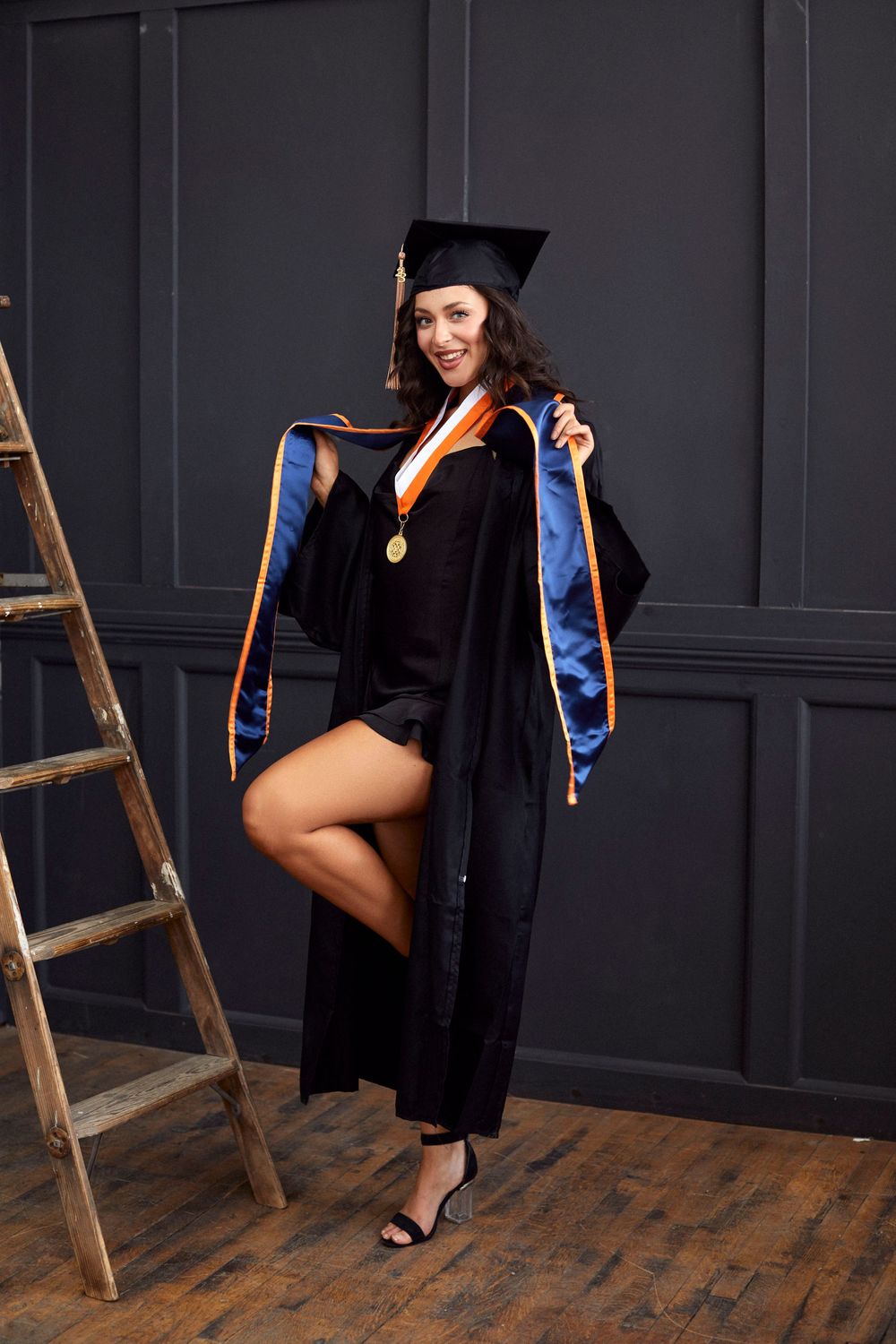 Graduate in academic regalia and blue sash poses with wooden ladder in studio.