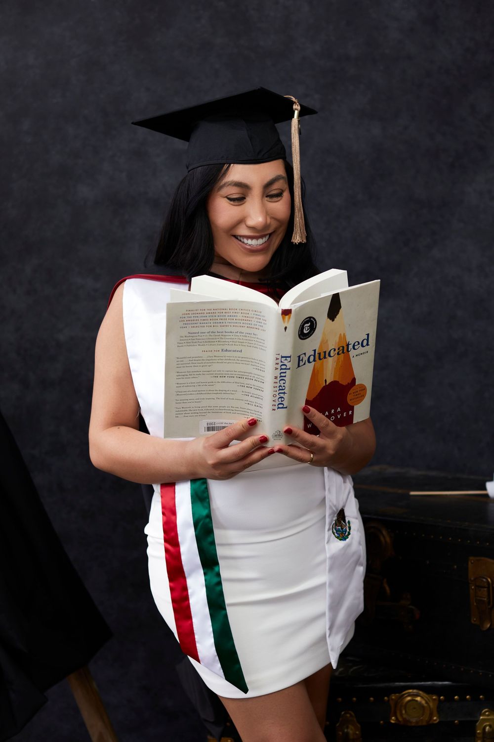 Graduate in white dress with Mexican flag colors reads a book against dark background.