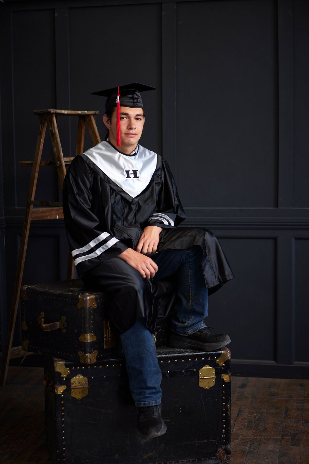 Graduate in black gown sits on wooden chest against dark wall in casual portrait.