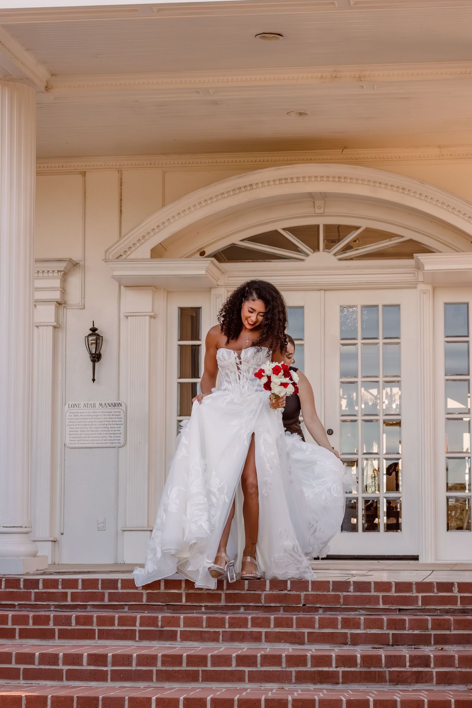 Bride in elegant white gown with thigh slit poses on brick steps of mansion with red flower bouquet.