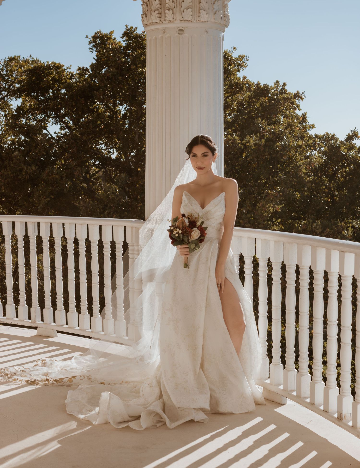 Bride in elegant white gown poses on sunlit balcony with classical architecture.
