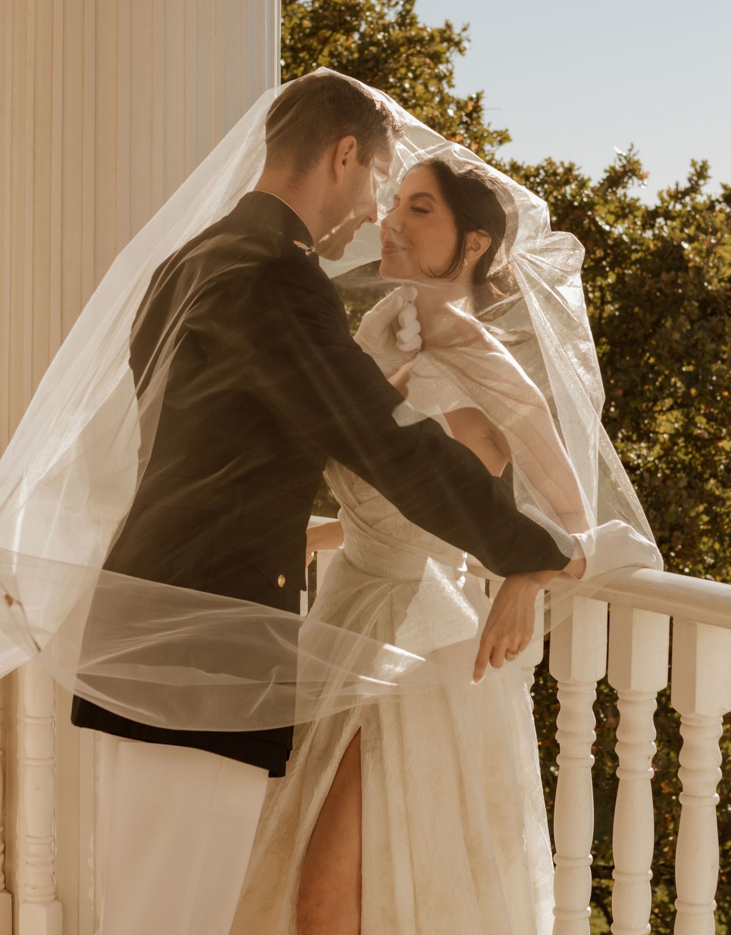 Romantic couple embraces on balcony with flowing veil in golden sunlight.