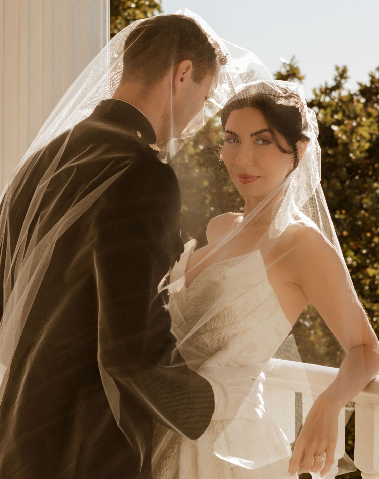 Romantic wedding portrait of couple in sunlight on a porch as veil flows in gentle breeze.