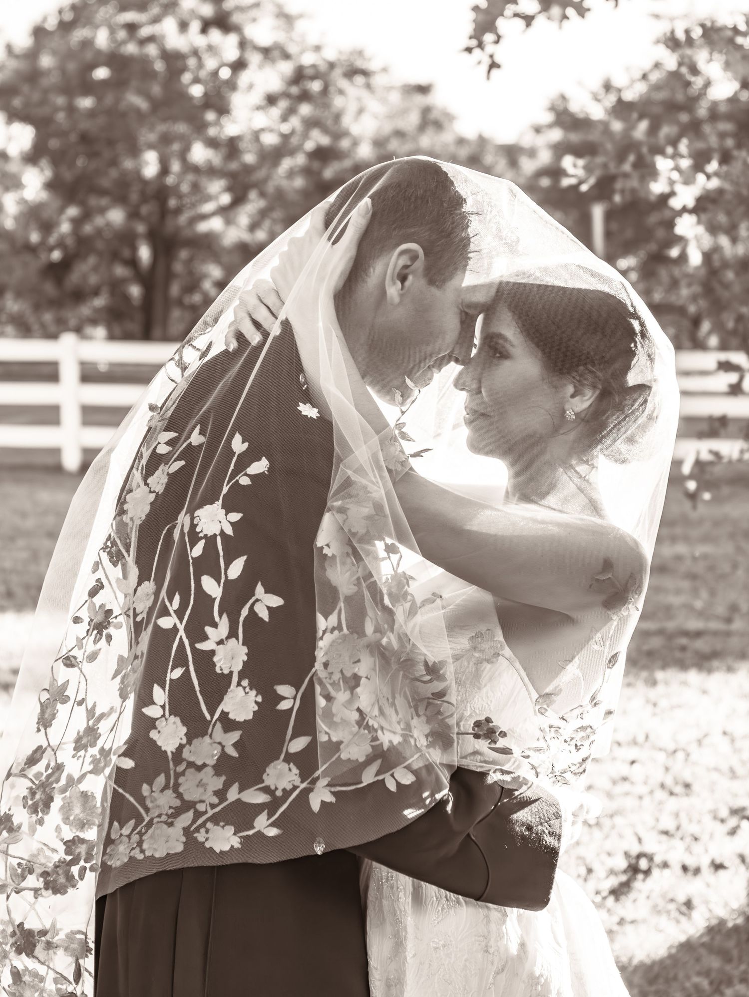 Black and white wedding photo with couple under floral embroidered veil outdoors.