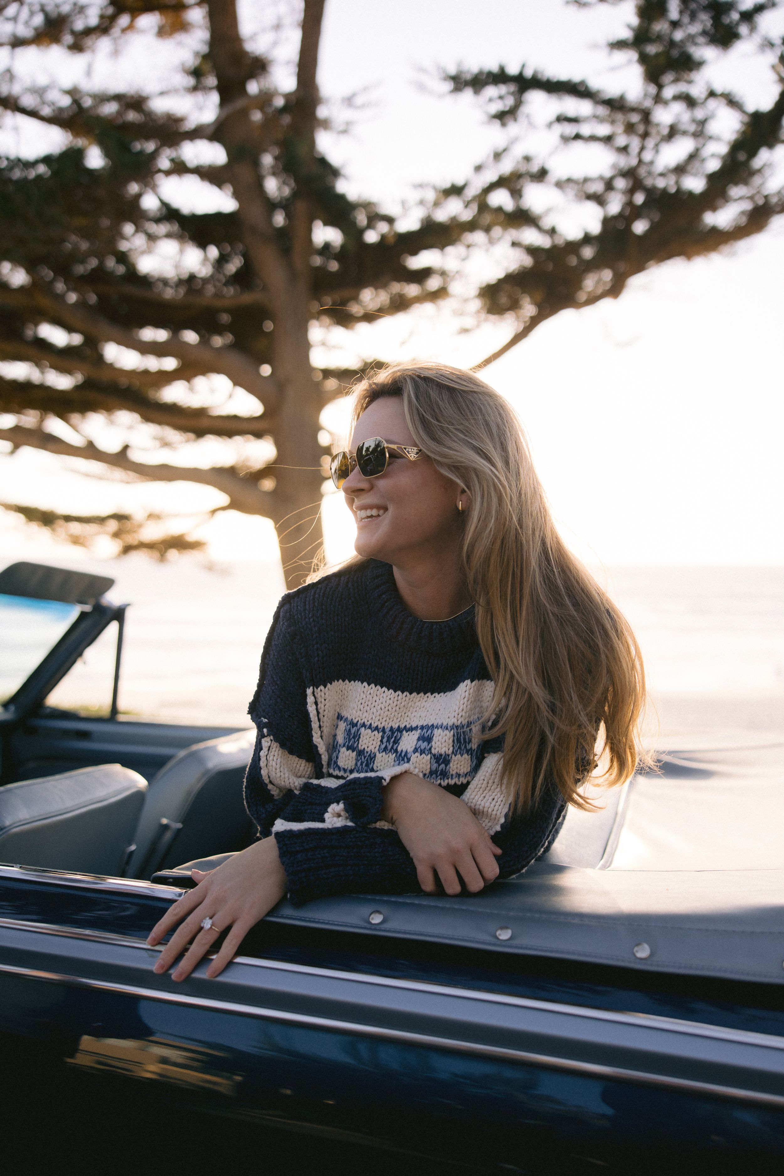 A person wearing sunglasses and a sweater leans out of a vintage car window during golden hour with trees in background.