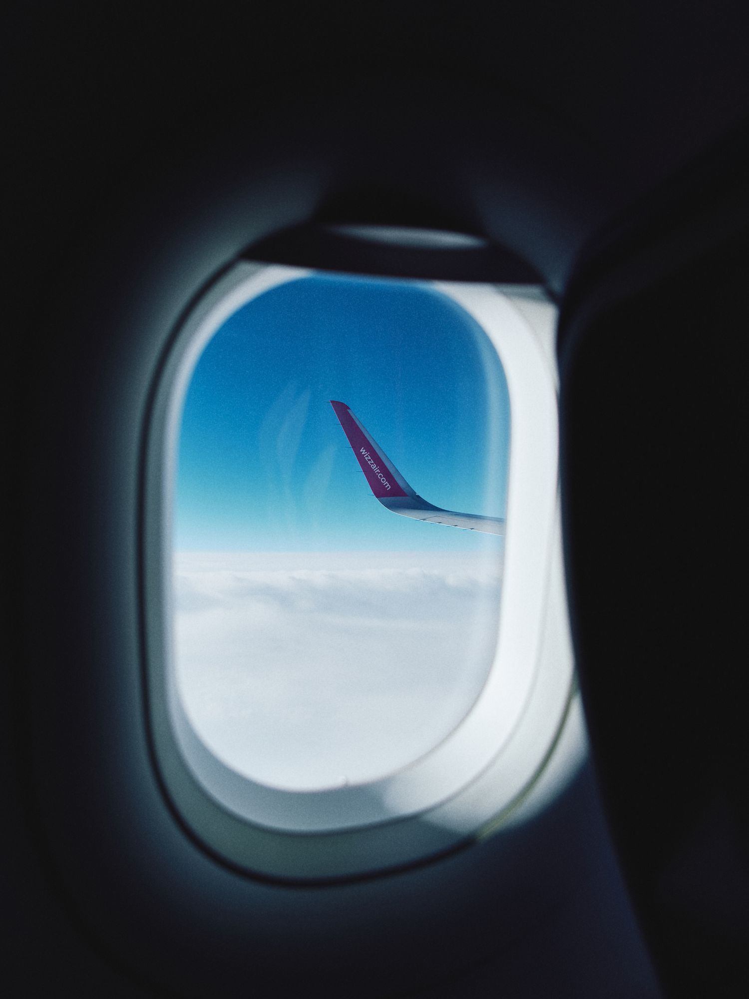View from airplane window showing a pink or magenta wing tip against blue sky and white clouds below.