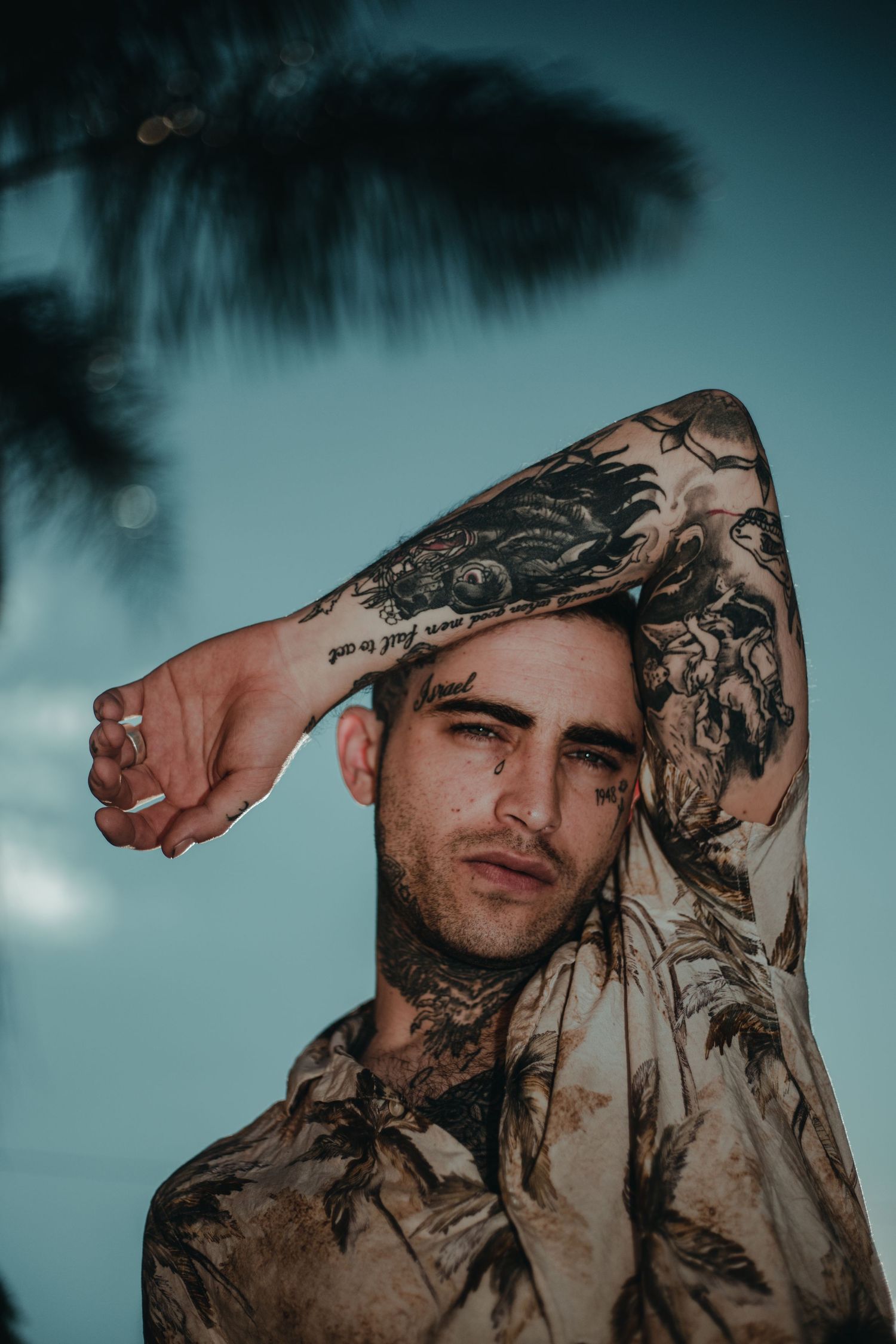 Artistic portrait with palm tree silhouette against turquoise sky showing tattooed arm raised overhead.